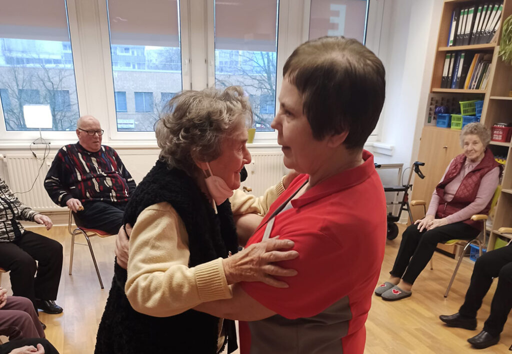 Pflegekraft umarmt &auml;ltere Frau in einem Gemeinschaftsraum mit Senioren.