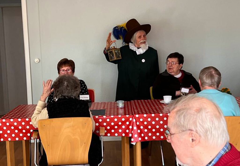 Ein &auml;lterer Herr in historischer Kleidung steht mit einer Laterne in der Hand neben einem Tisch, an dem Senioren sitzen.