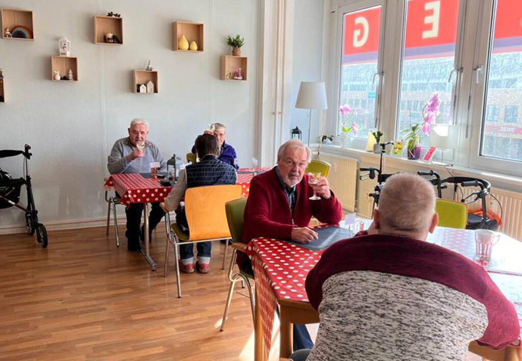 Ältere Menschen in einem Gemeinschaftsraum, sitzen an Tischen mit rot-weiß gepunkteten Tischtüchern.