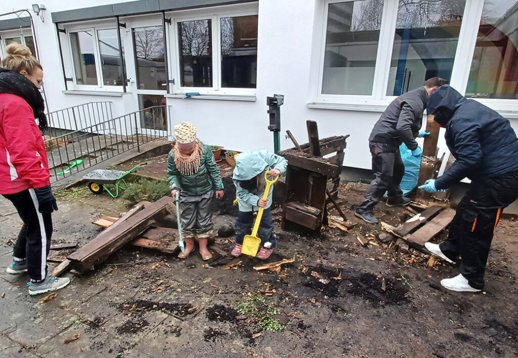 Personengruppe, darunter zwei Kinder, beim Arbeiten im Freien vor einem Geb&auml;ude.
