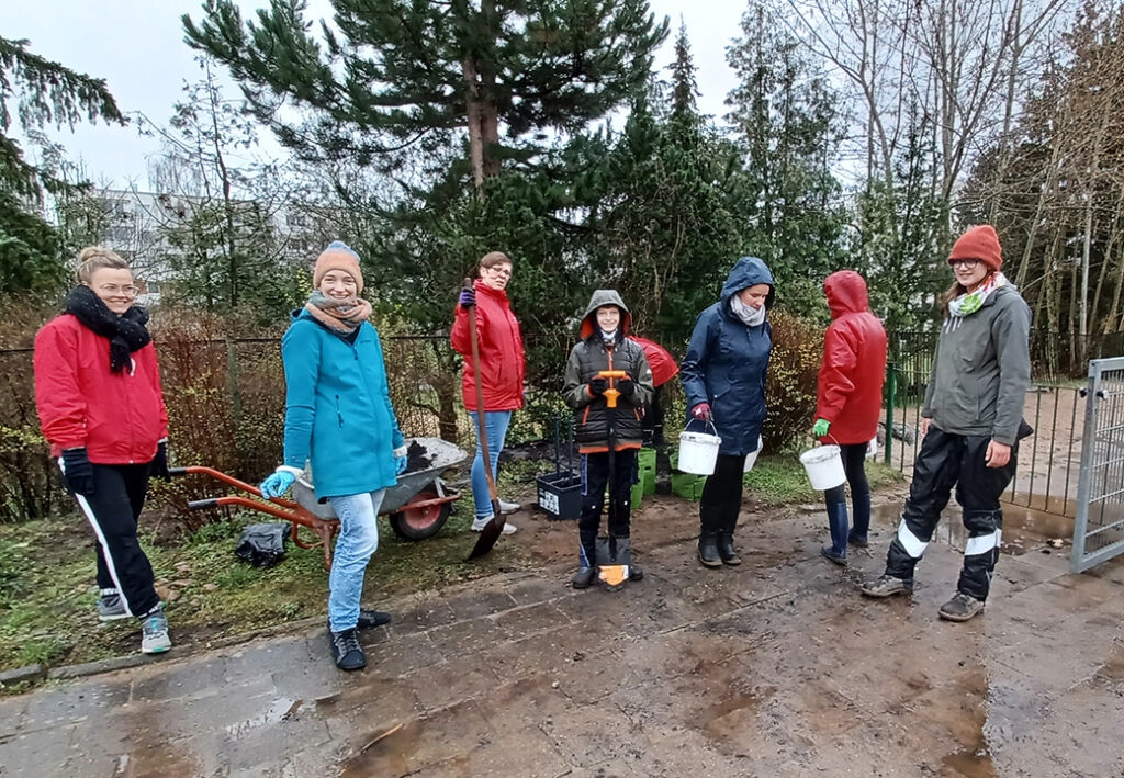 Eine Gruppe von Menschen steht drau&szlig;en in Regenkleidung und mit Gartenger&auml;ten.