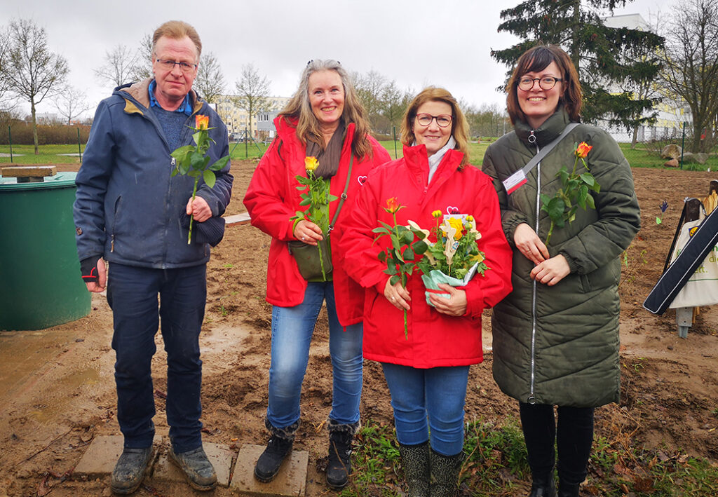 Vier Personen stehen nebeneinander in einem Freien, Gartenarbeit wird angedeutet.