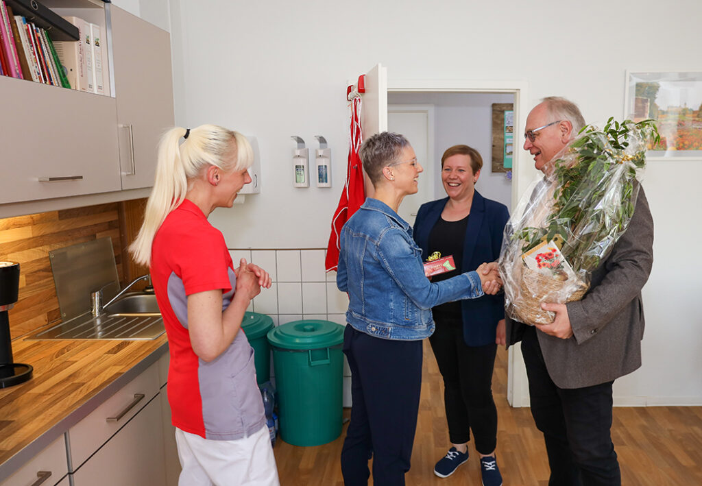 Personengruppe in einem Raum, eine Frau schüttelt die Hand eines Mannes mit einem eingepackten Geschenk.