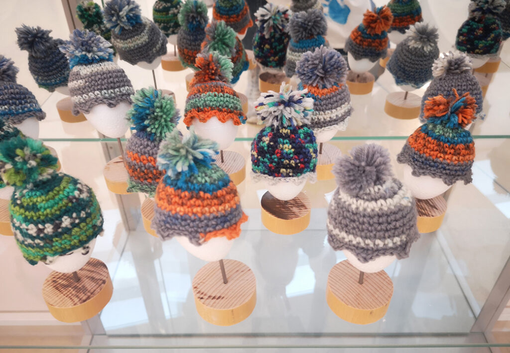 Eine Ausstellung von kleinen Strickm&uuml;tzen mit farbigen Bommeln auf wei&szlig;en Styropork&ouml;pfen, die auf Holzst&auml;ndern stehen.
