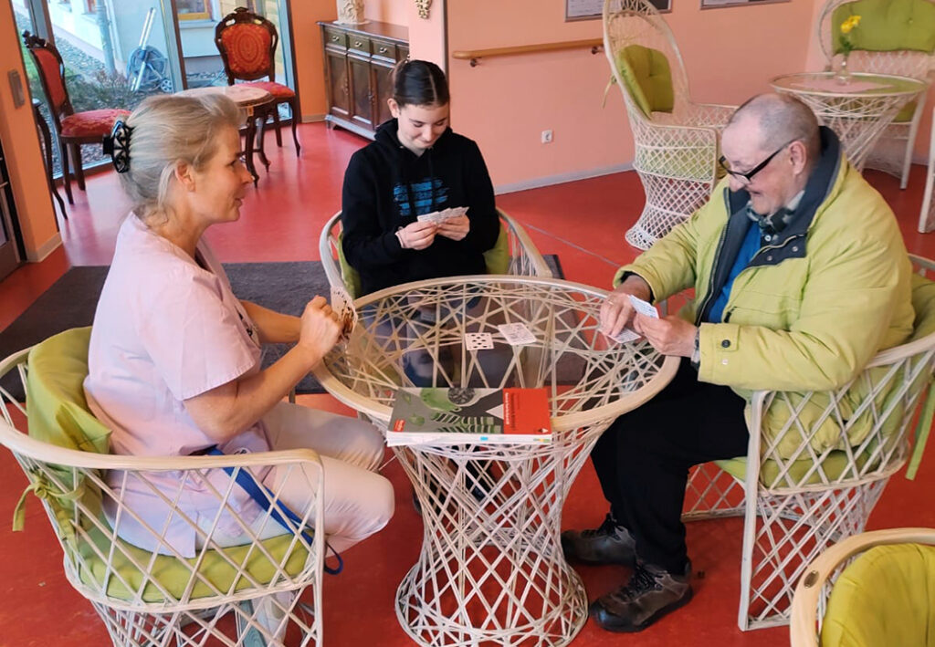 Drei Personen spielen Karten an einem Tisch in einem Gemeinschaftsraum.