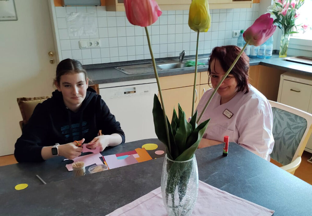 Zwei Personen sitzen an einem Tisch und basteln mit Papier, w&auml;hrend Tulpen in einer Vase auf dem Tisch stehen.
