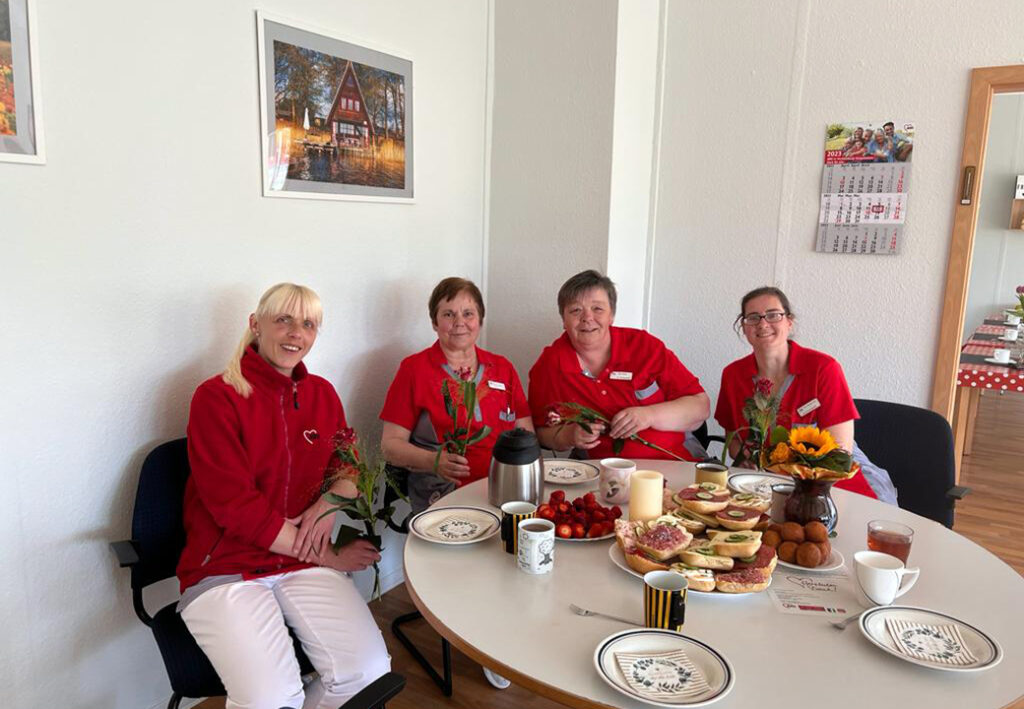 Vier Frauen in roten Uniformen sitzen l&auml;chelnd an einem Tisch mit Essen.