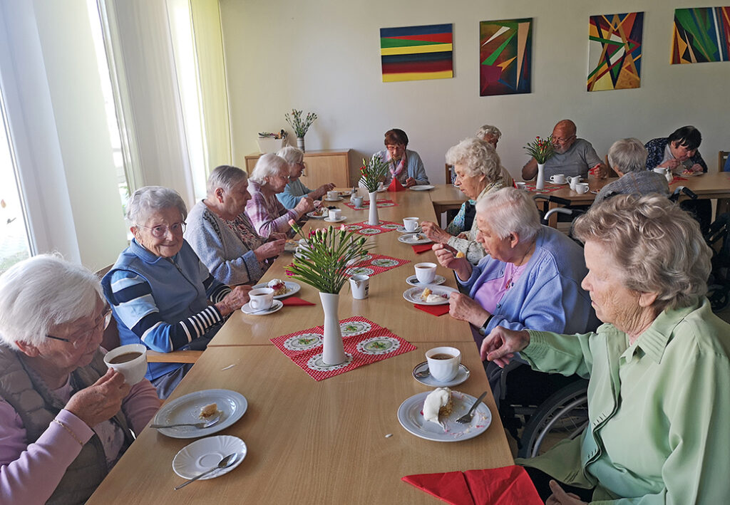 Eine Gruppe älterer Menschen sitzt an einem langen Tisch und trinkt Tee oder Kaffee.