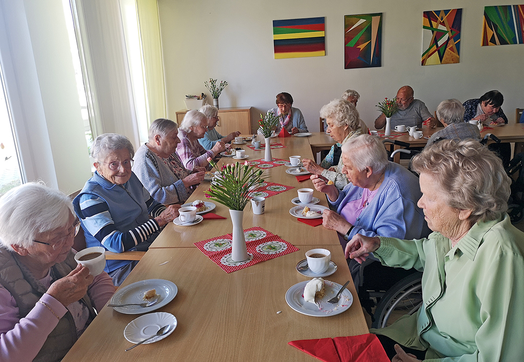 Eine Gruppe älterer Menschen sitzt an einem langen Tisch und trinkt Tee oder Kaffee.
