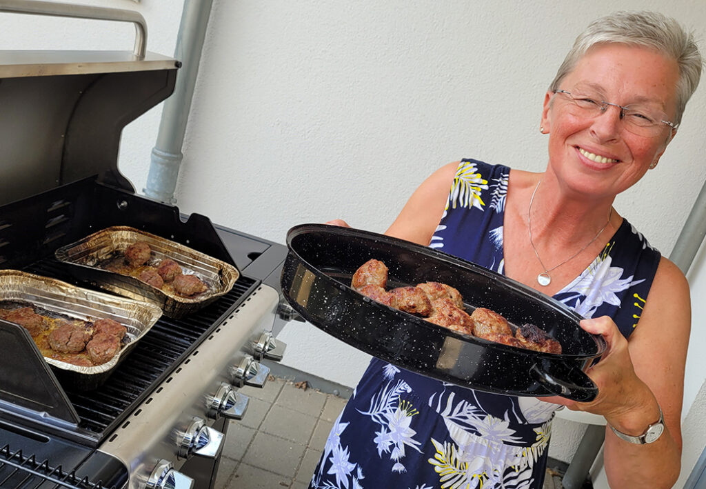 Frau hält Backblech mit Fleischklößchen neben einem Grill