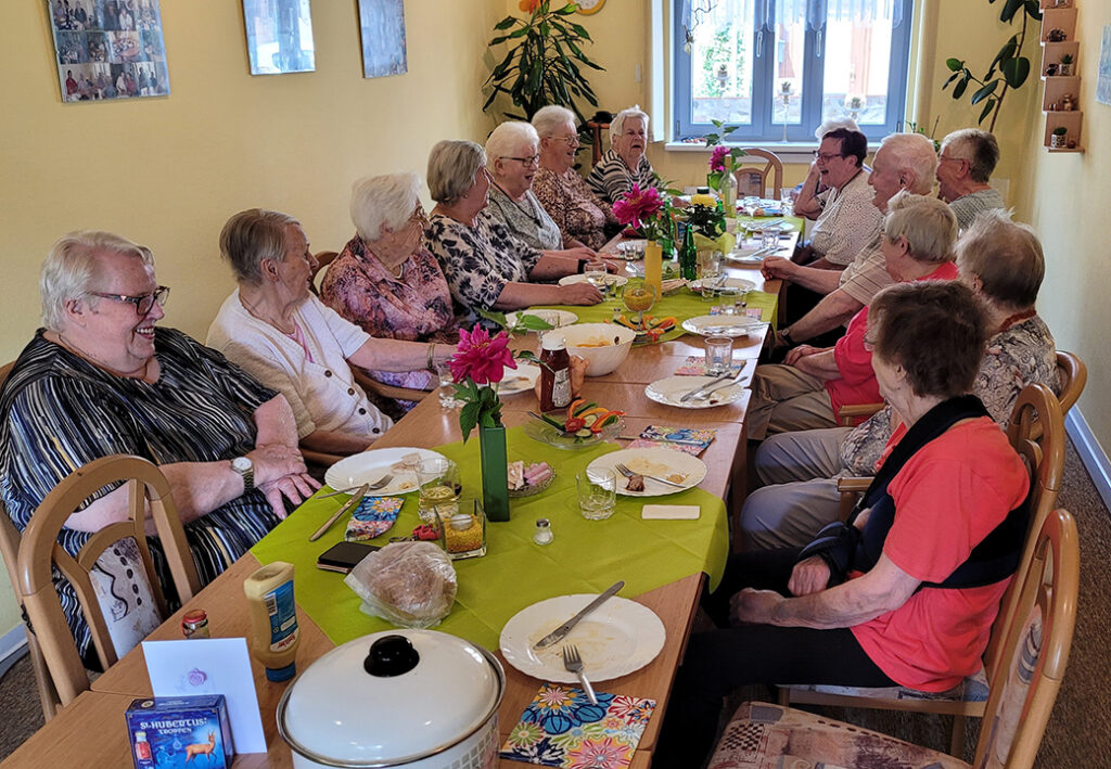 Eine Gruppe &auml;lterer Menschen sitzt gemeinsam an einem langen Tisch, fr&ouml;hlich beim Essen und Plaudern.