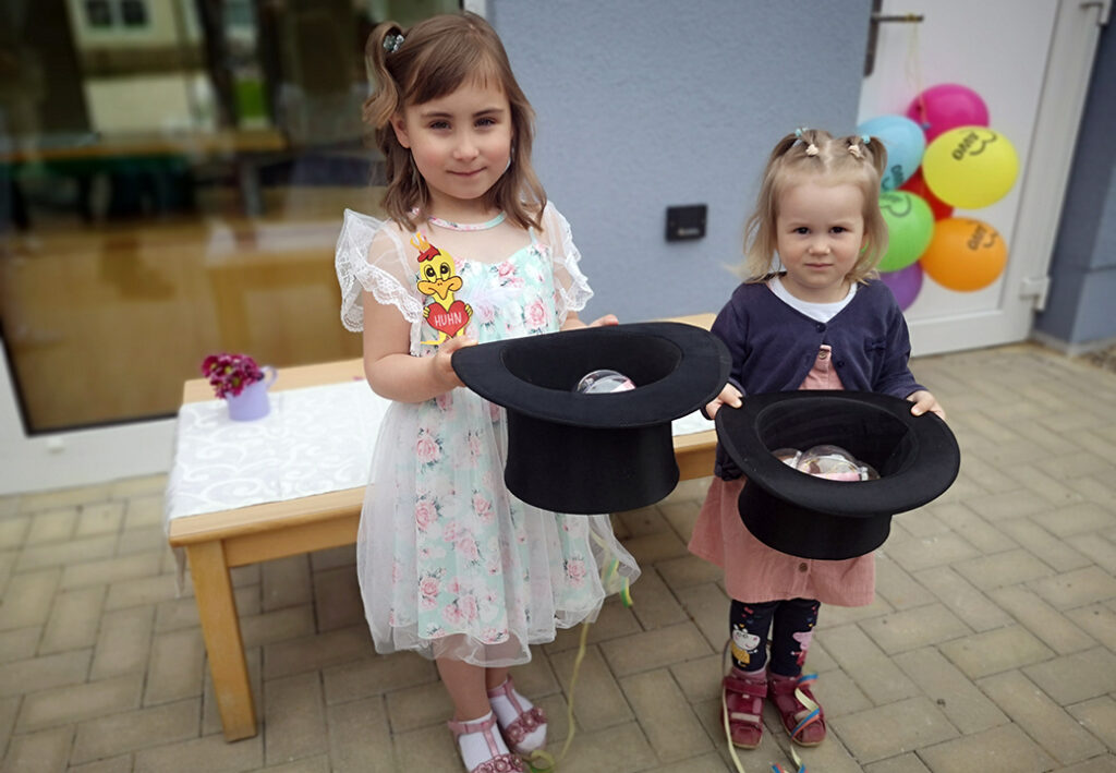 Zwei kleine M&auml;dchen halten Zauberh&uuml;te und stehen vor einem Tisch mit Ballons im Hintergrund.