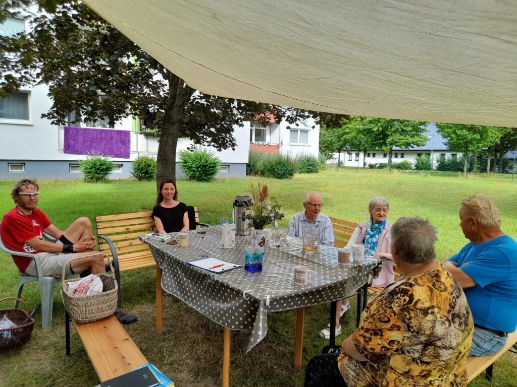 Mehrere Personen sitzen an einem Tisch unter einem Sonnensegel im Freien.