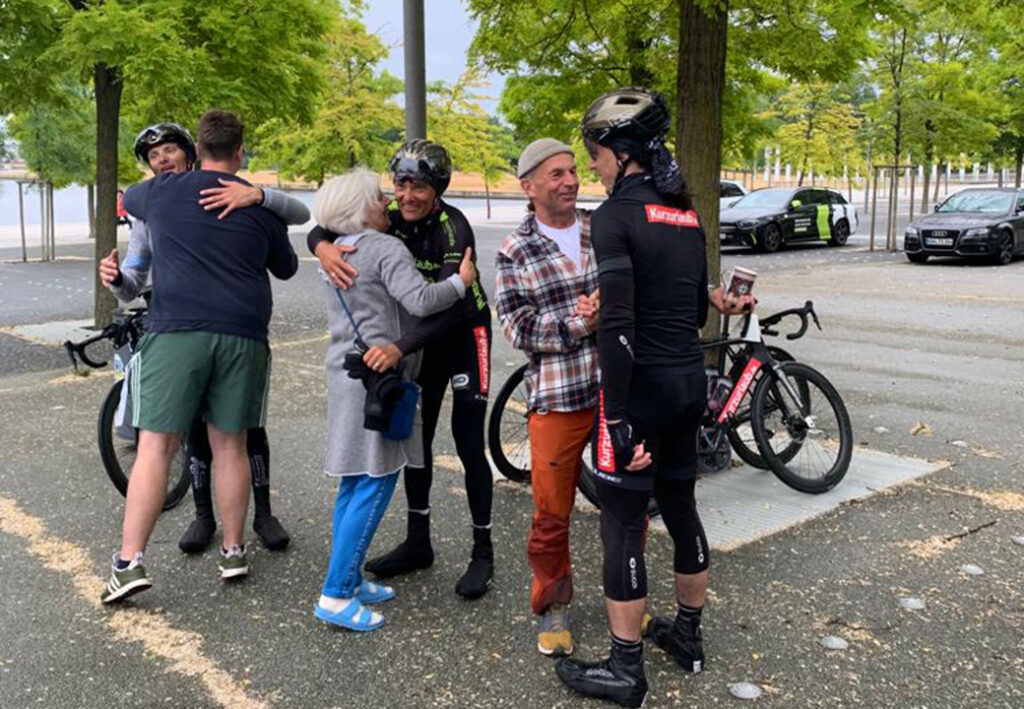 Eine Gruppe von Menschen, darunter Radfahrer in schwarzer Sportbekleidung und Helmen, die sich auf einem Fahrradweg umarmen und miteinander sprechen.