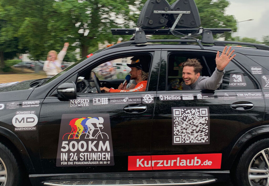 Ein schwarz lackiertes Auto mit Aufklebern und Logos verschiedener Sponsoren. Zwei Personen sind im Auto sichtbar, eine winkt aus dem Fenster. Ein gro&szlig;er Aufkleber mit der Aufschrift '500KM IN 24 STUNDEN F&Uuml;R DIE FRAUENH&Auml;USER IN M-V' ist auf der Seite zu sehen.