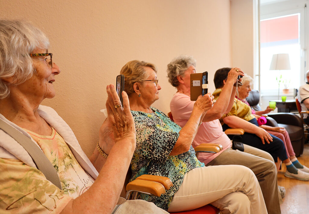 Eine Gruppe älterer Frauen sitzt in einer Reihe auf Stühlen und hält Smartphones oder Tablets in den Händen.