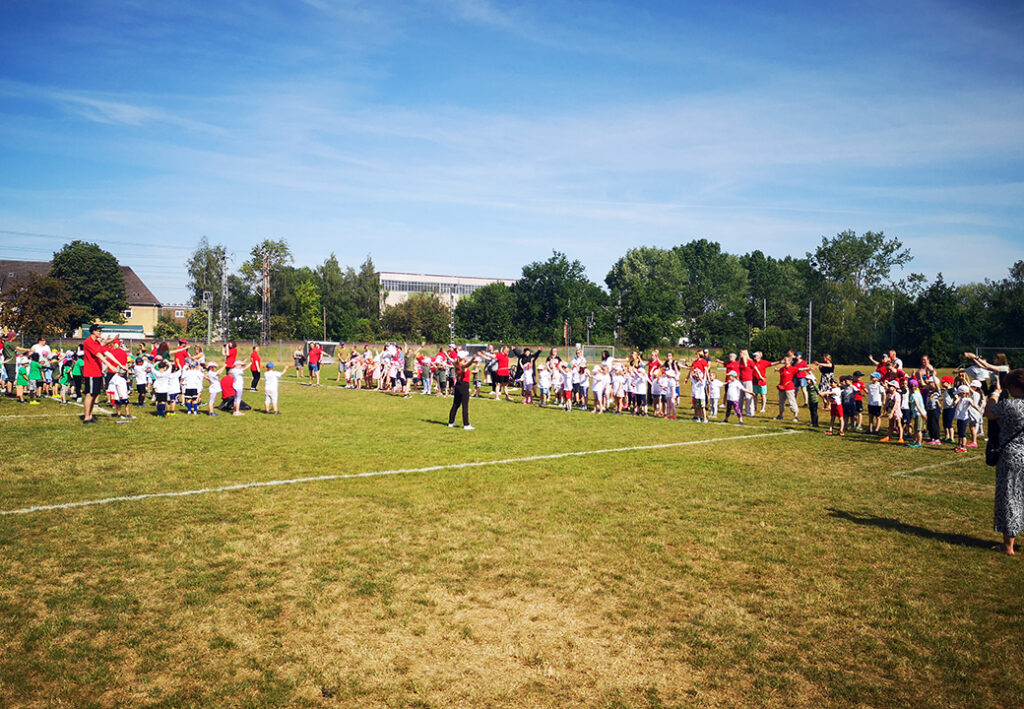 Mehrere Gruppen von Kindern und Erwachsenen stehen auf einem Sportplatz.