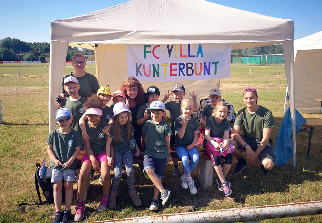 Kinder und Betreuer unter einem Zelt mit einem Banner 'FC Villa Kunterbunt'.