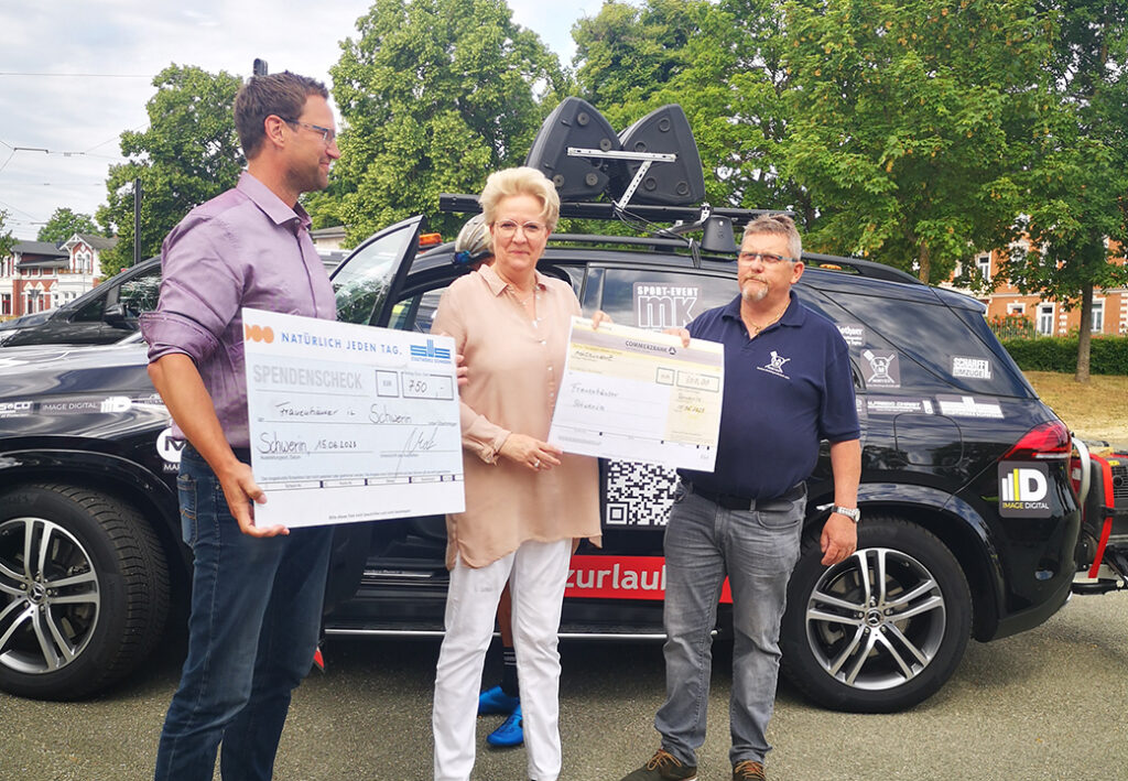 Drei Personen posieren mit Spendenschecks vor einem Auto.