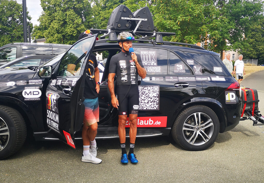 Zwei M&auml;nner in Sportkleidung vor einem schwarzen Fahrzeug mit Aufklebern