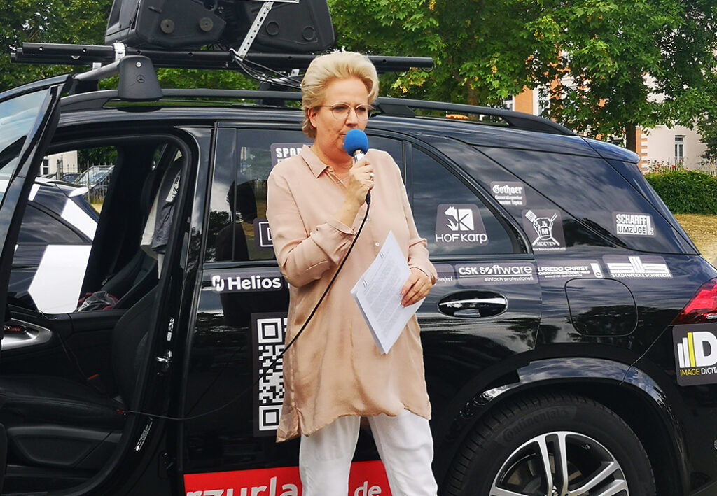 Eine Frau mit kurzem, blondem Haar steht vor einem schwarzen Auto mit offenen T&uuml;ren und spricht in ein blaues Mikrofon. Sie h&auml;lt ein Blatt Papier in der anderen Hand. Das Auto ist mit diversen Aufklebern und einem Dachtr&auml;ger ausgestattet.