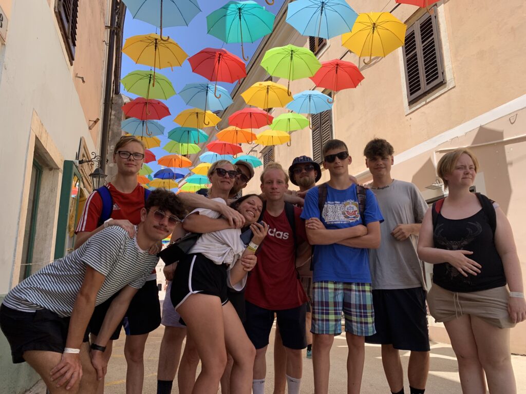 Gruppenfoto einer fr&ouml;hlichen Gruppe von Personen in einer schmalen Gasse mit bunten Regenschirmen an der Decke.