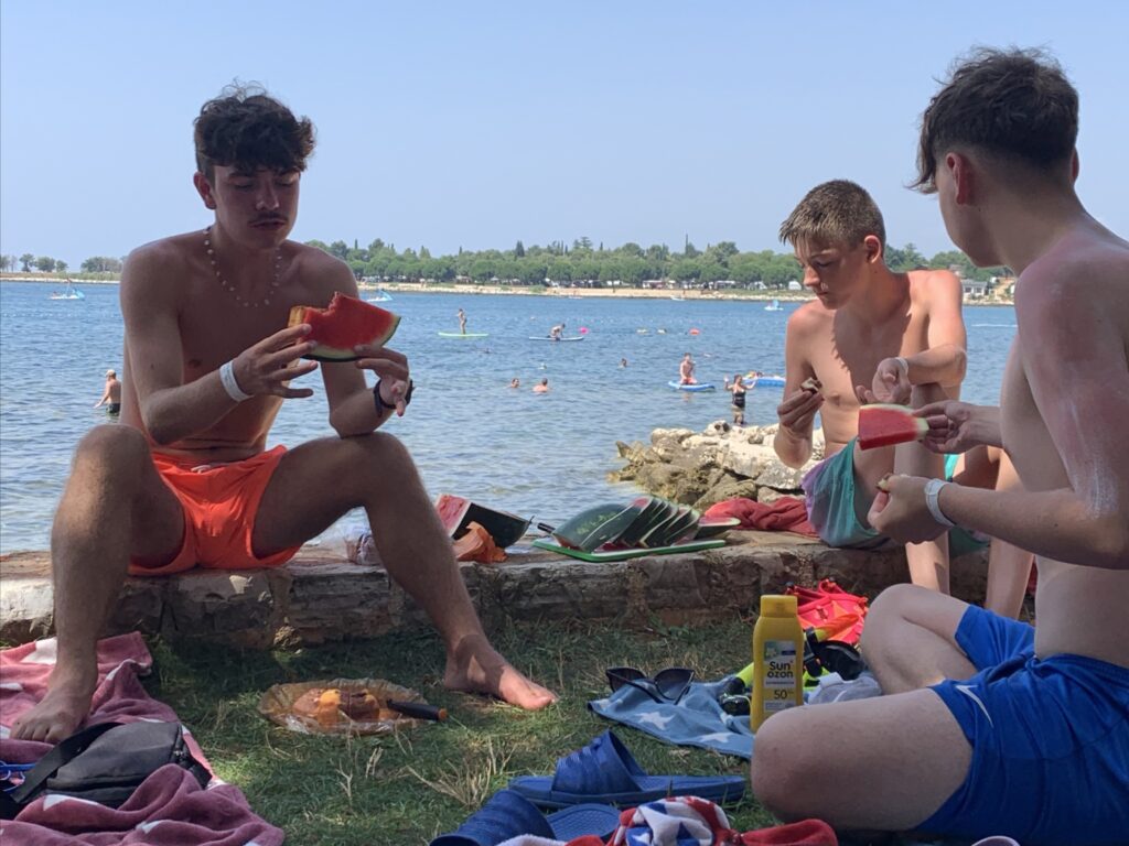 Drei junge M&auml;nner essen Wassermelone am Strand.