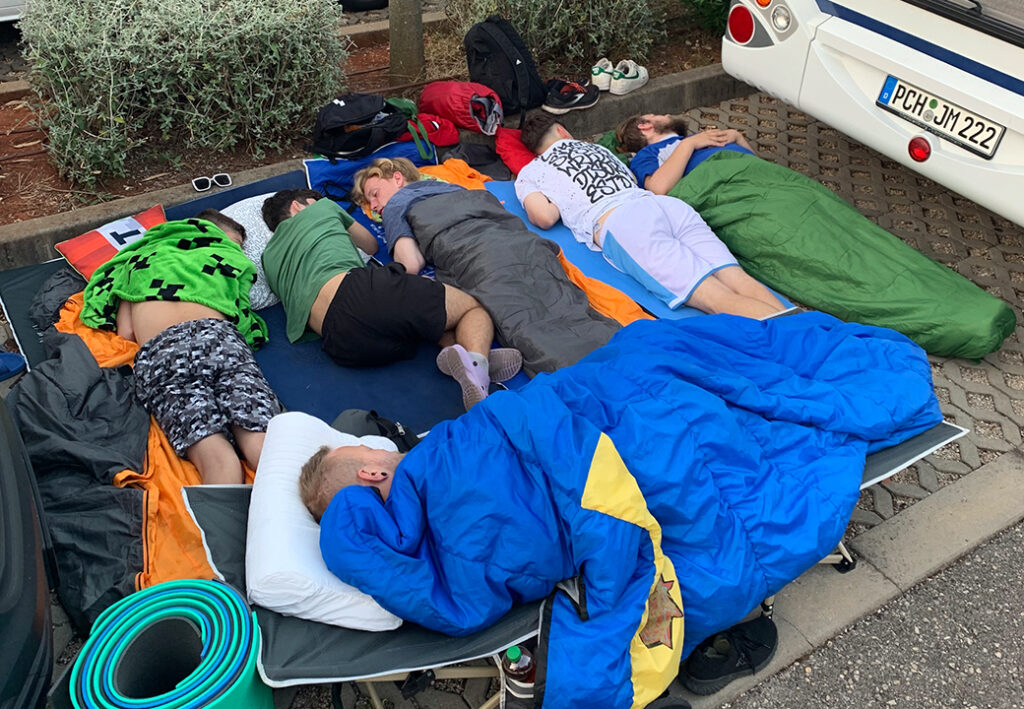 F&uuml;nf Personen in Schlafs&auml;cken auf Campingmatten schlafen nebeneinander im Freien neben einem Fahrzeug.