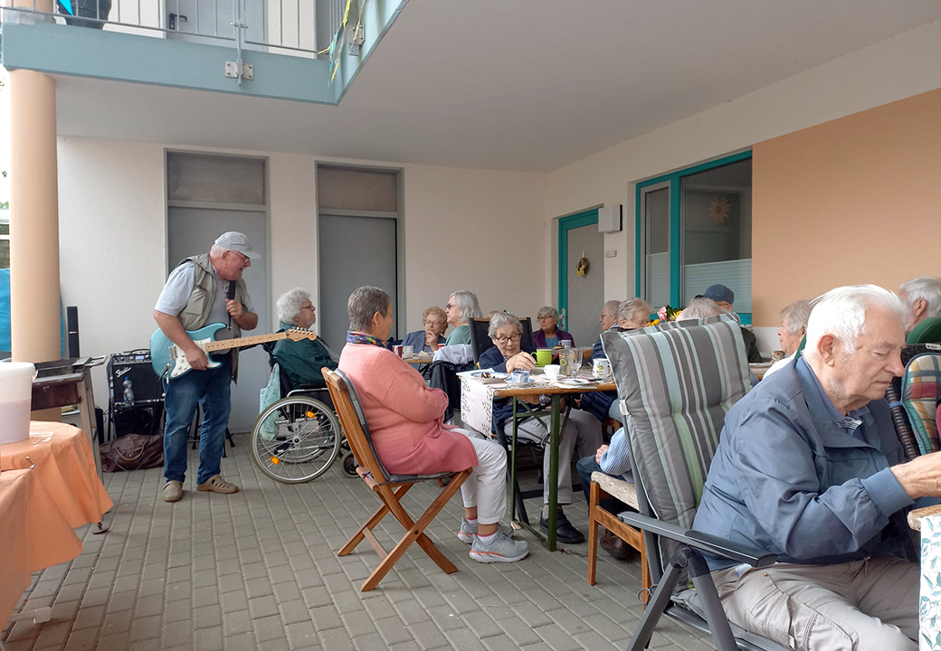 Eine Gruppe älterer Menschen bei einem sommerlichen Treffen im Freien, ein Mann spielt Gitarre.