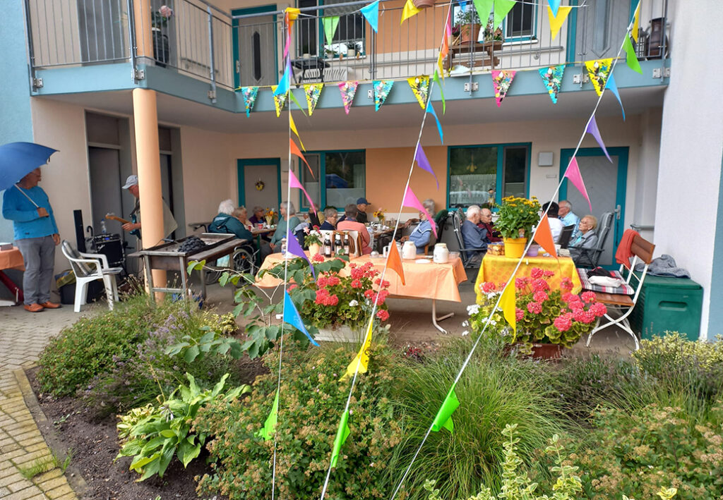 Ein Gemeinschaftsbereich mit Menschen, die an Tischen im Freien sitzen, umgeben von farbenfrohen Pflanzen und Wimpeln.