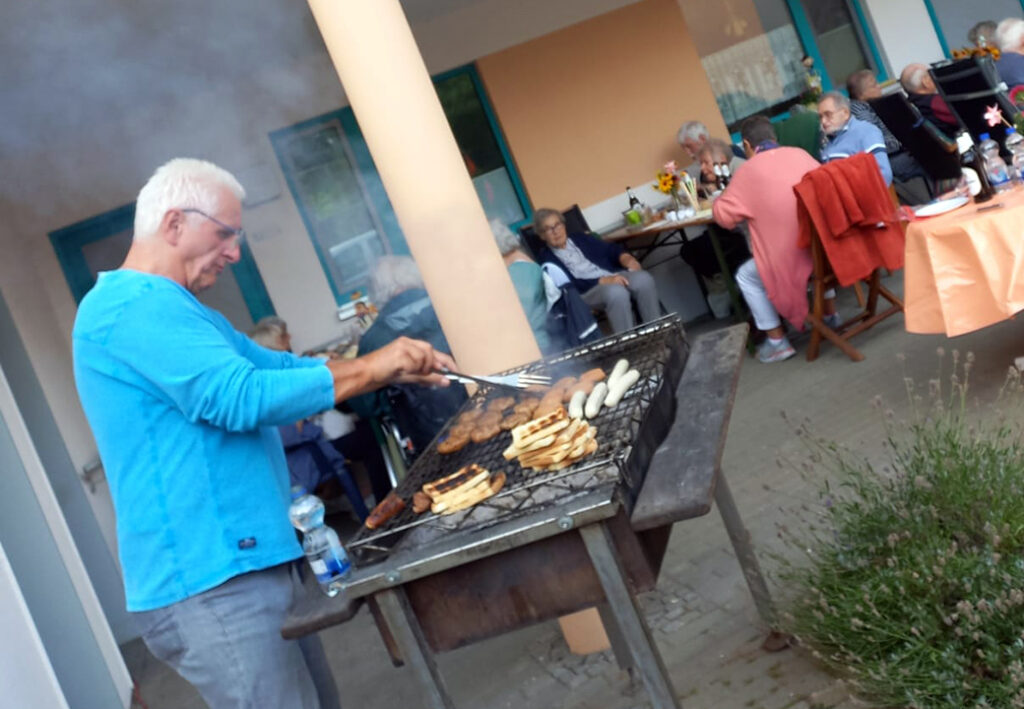 Eine Person grillt Essen auf einem Grill, w&auml;hrend im Hintergrund Menschen um einen Tisch sitzen.