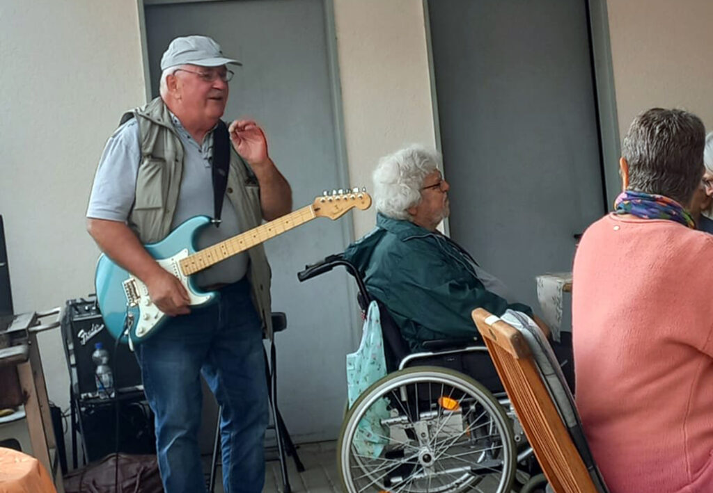 Ein Mann spielt Gitarre neben einer sitzenden Person im Rollstuhl und einer weiteren sitzenden Person mit einem Schal.