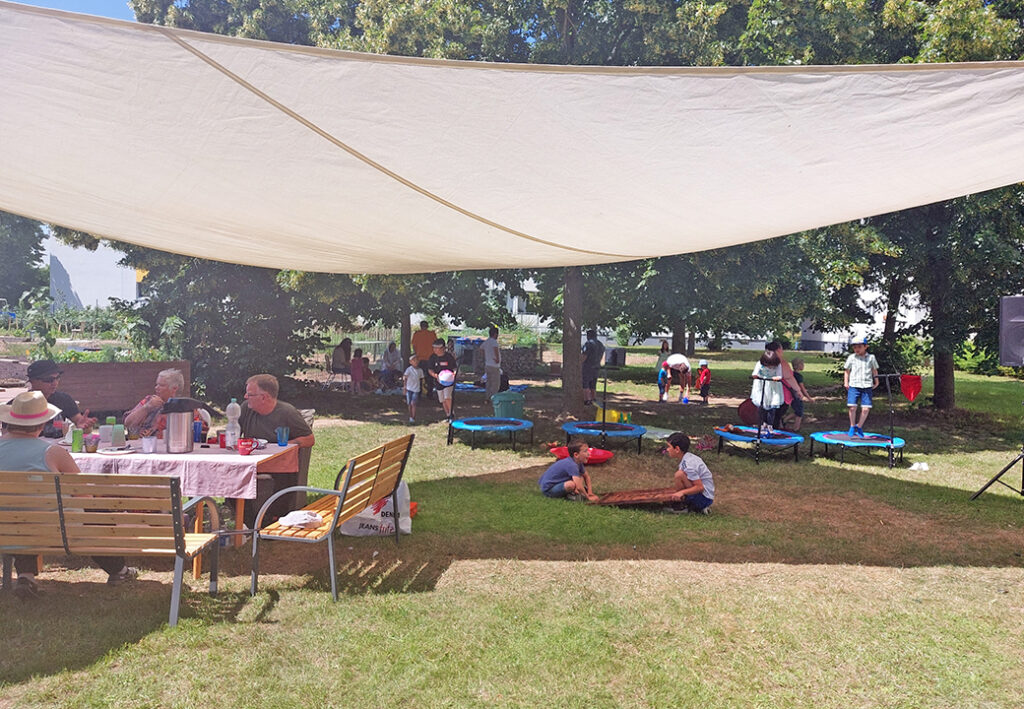 Menschen bei einer Veranstaltung im Freien, unter einem gro&szlig;en Sonnensegel sitzend und spielend.