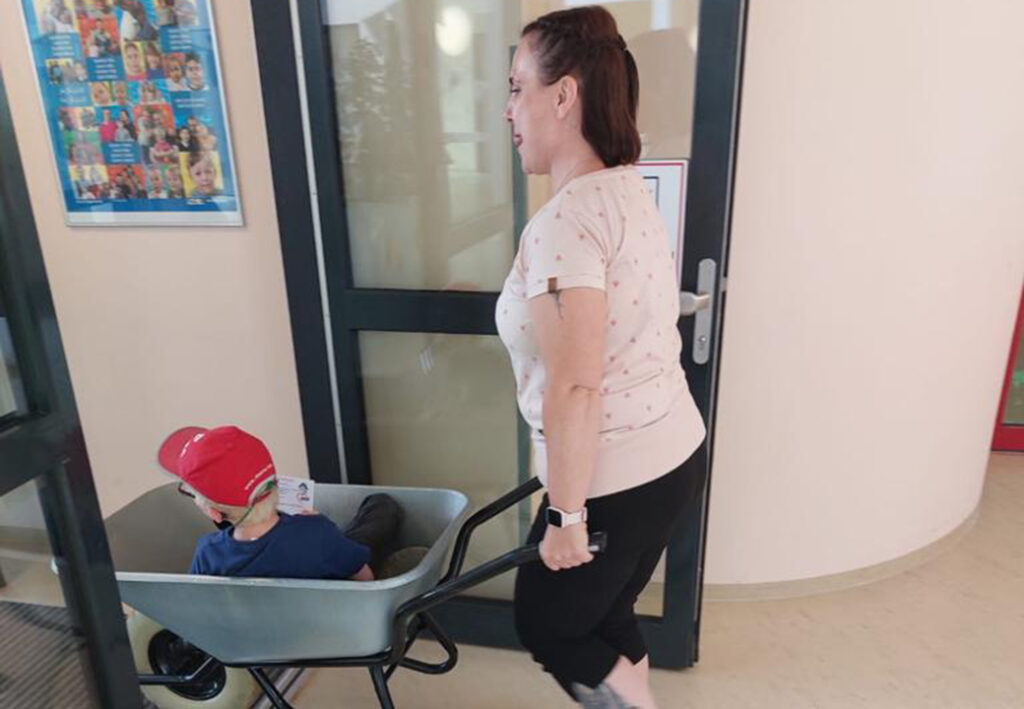 Eine Frau schiebt in einem Geb&auml;ude einen kleinen Jungen, der in einer Schubkarre sitzt.