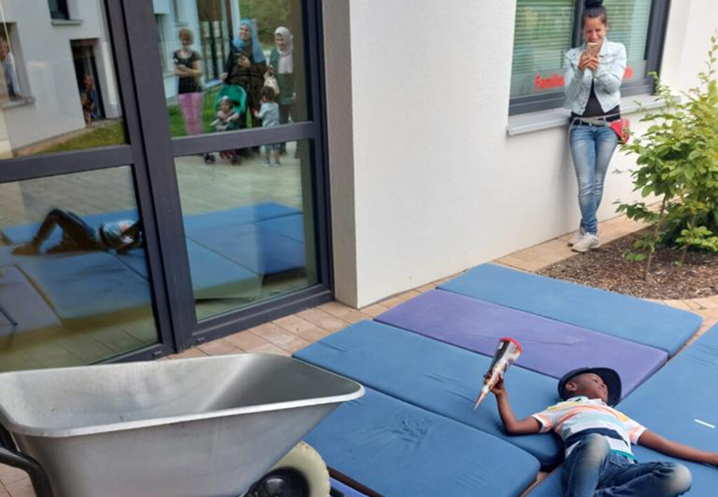 Kind liegt auf Matten mit einem Spielzeug im Hof neben einer Schubkarre, w&auml;hrend eine Frau ihn beobachtet und fotografiert.