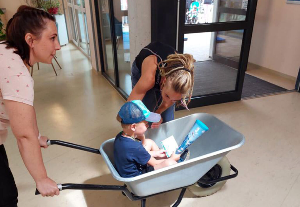 Eine Frau schiebt eine Schubkarre mit einem sitzenden Kind darin, w&auml;hrend eine andere Frau sich &uuml;ber das Kind beugt.
