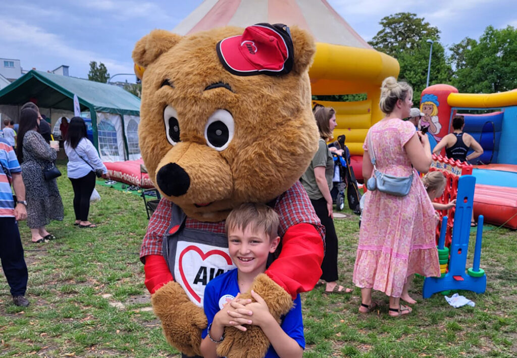 Ein Kind umarmt einen gro&szlig;en Pl&uuml;schb&auml;ren mit AWO-Logo bei einer Outdoor-Veranstaltung.