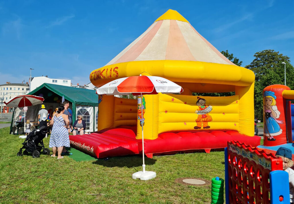 Eine aufblasbare H&uuml;pfburg in Form eines gelben Zirkuszelts mit rotem Boden steht auf einer gr&uuml;nen Wiese.