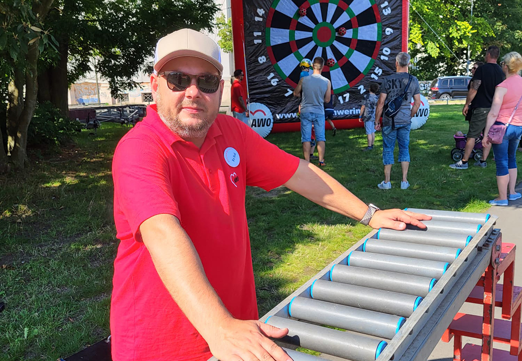 Ein Mann in rotem AWO-Polo bei einem Outdoor-Event, im Hintergrund eine große Dart-Scheibe und weitere Personen.