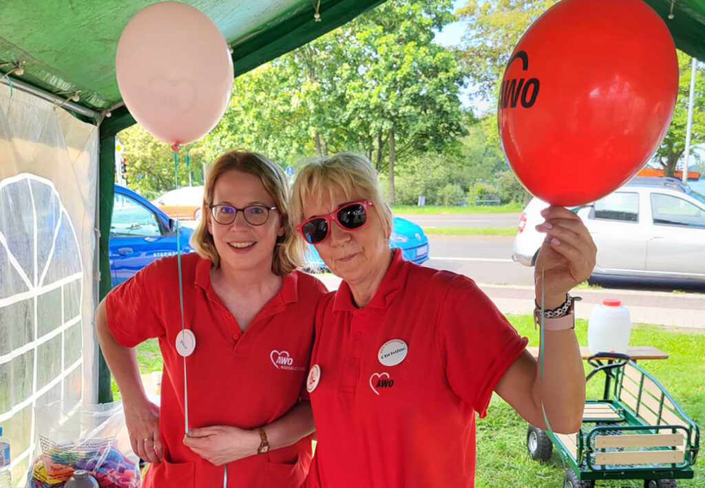 Zwei Frauen in roten AWO-Poloshirts stehen in einem Freiluftzelt und halten jeweils einen Luftballon.