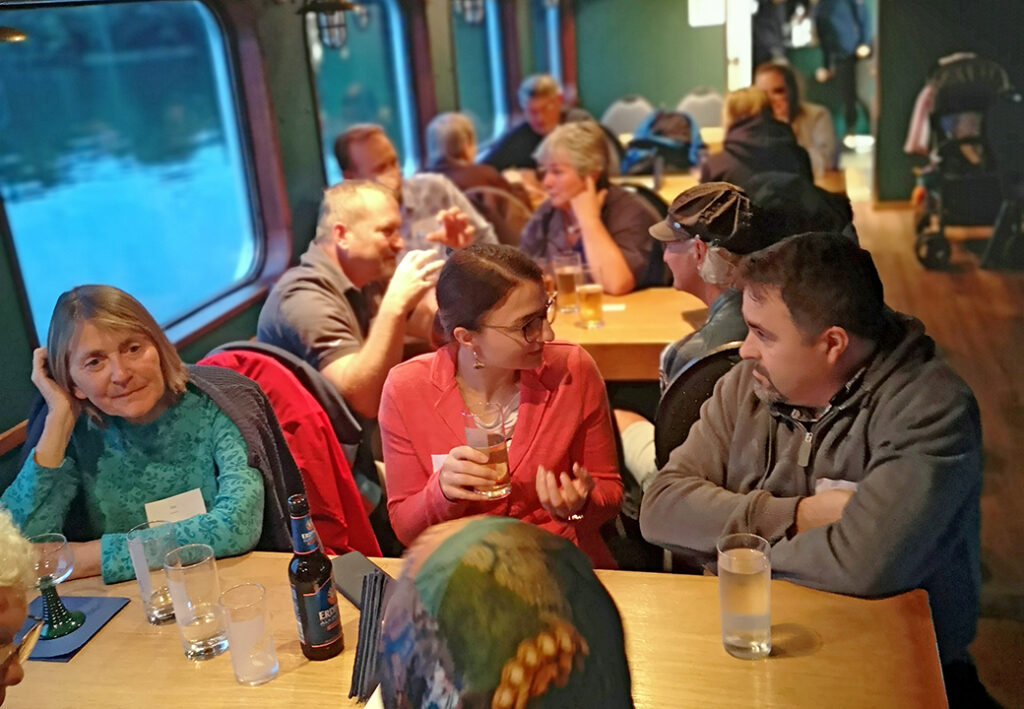 Eine Gruppe von Menschen sitzt in einem gem&uuml;tlichen Raum an langen Holztischen und unterh&auml;lt sich.