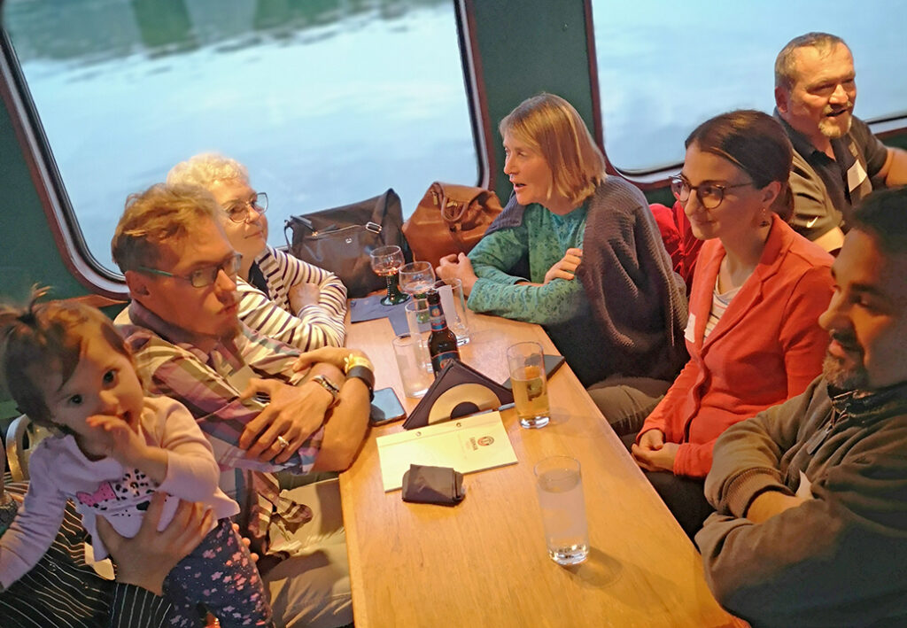 Eine Gruppe von Menschen sitzt in einem geselligen Kreis an einem Holztisch in einem Boot oder Zug mit gro&szlig;en Fenstern im Hintergrund.