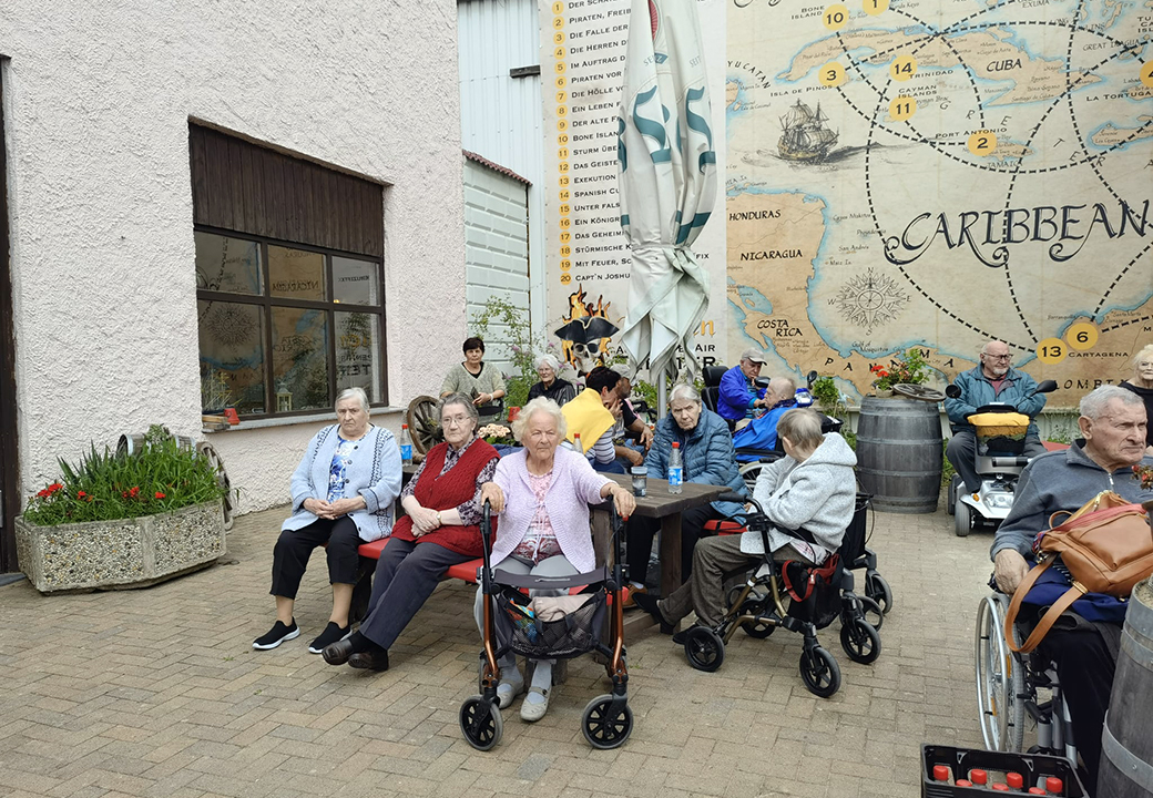 Eine Gruppe älterer Menschen sitzt draußen vor einem pastellfarbenen Gebäude.