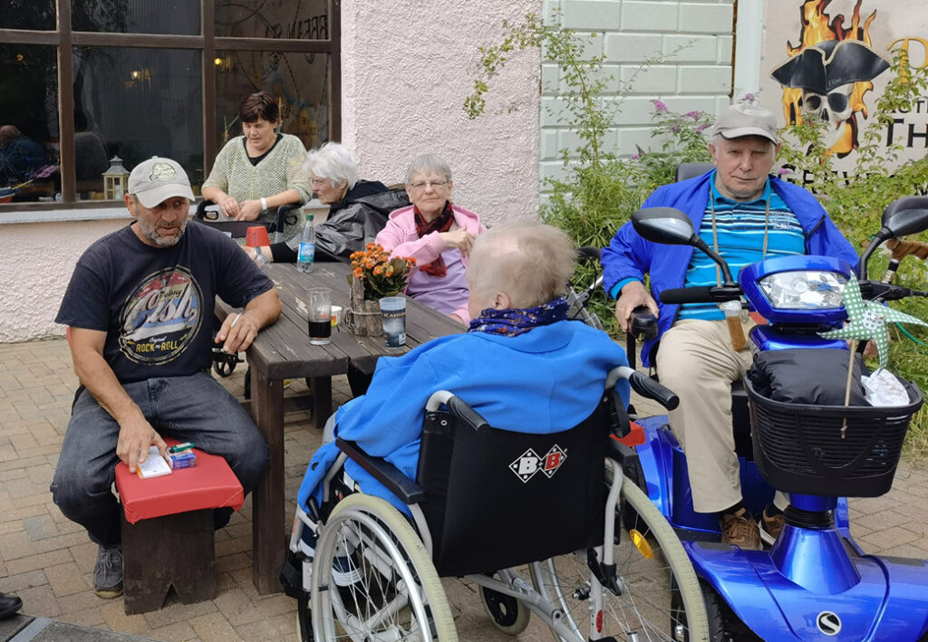 Eine Gruppe von Menschen sitzt drau&szlig;en an einem Tisch, darunter zwei Personen in Rollst&uuml;hlen.