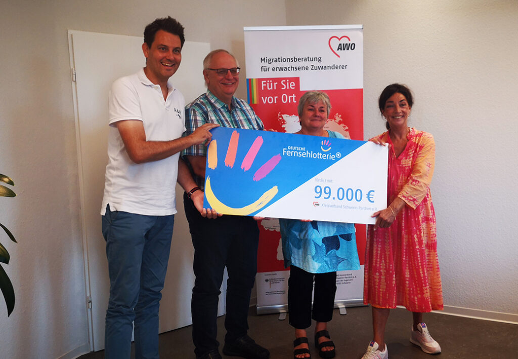 Vier Personen halten einen großen symbolischen Scheck über 99.000 Euro in einem Innenraum.