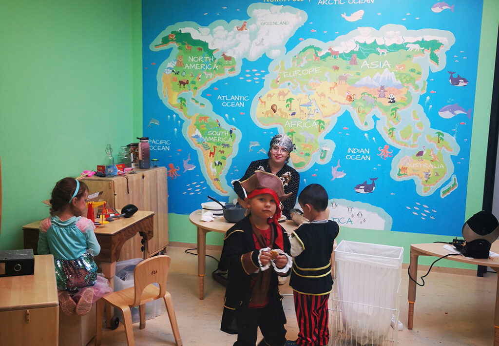 Kinder und eine Erzieherin in einem Raum mit einer gro&szlig;en Weltkarte an der Wand.