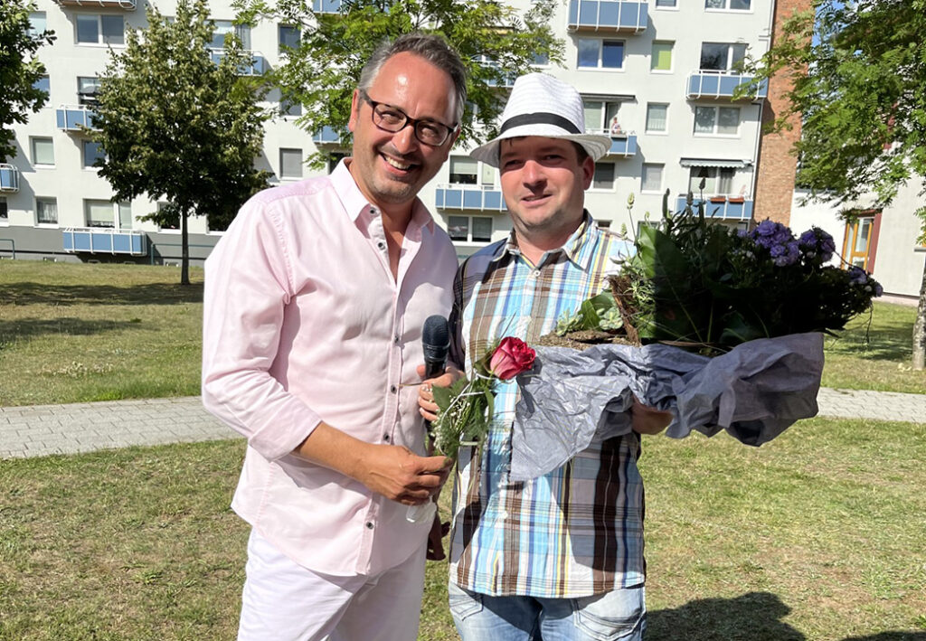 Zwei M&auml;nner stehen im Freien, einer h&auml;lt Blumen und der andere ein Mikrofon.