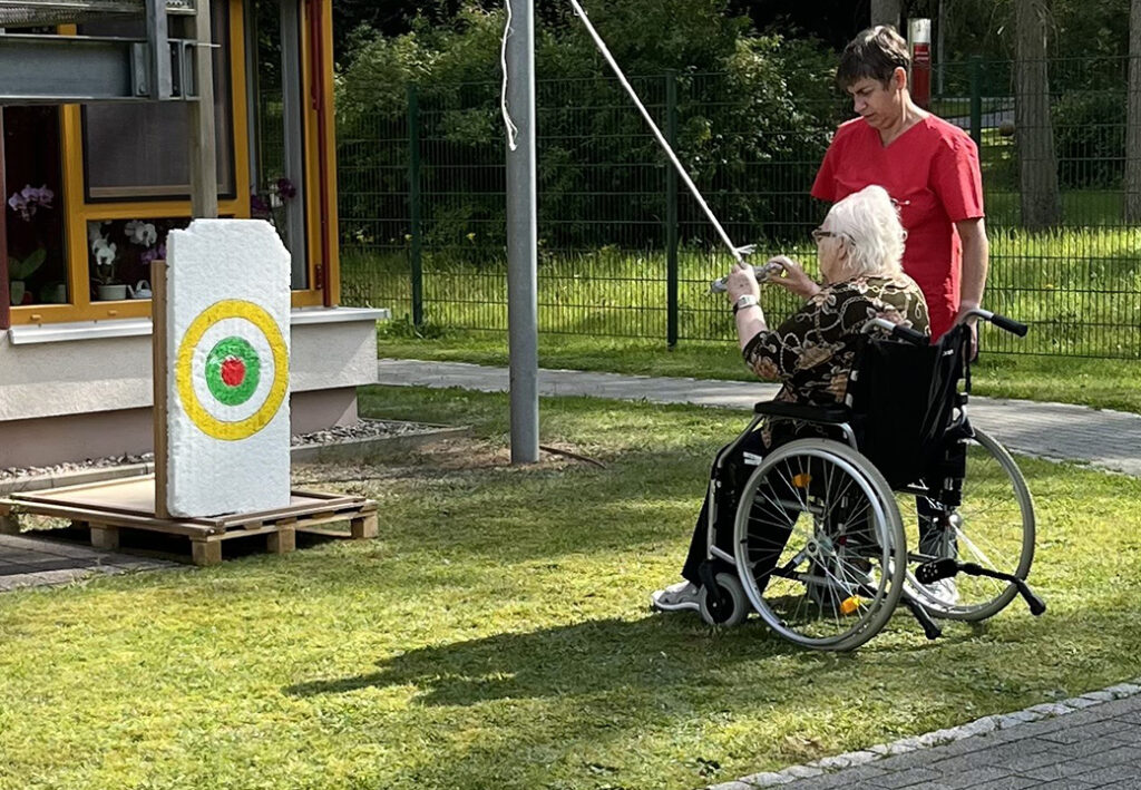 Eine &auml;ltere Person im Rollstuhl wird von einer Betreuungsperson beim Bogenschie&szlig;en im Freien unterst&uuml;tzt.
