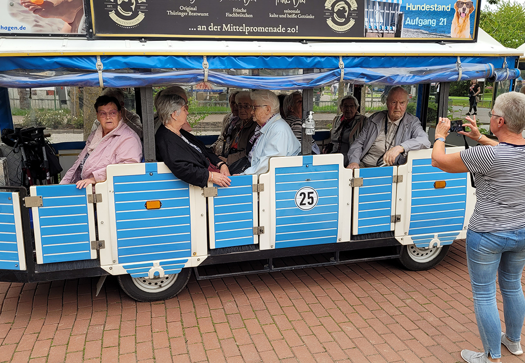 Eine Gruppe älterer Menschen sitzt in einer blau-weißen, offenen Bahn auf einem gepflasterten Weg. Eine Frau steht außen und fotografiert die Gruppe.