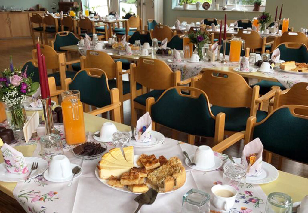 Gedeckter Tisch in einem Gemeinschaftsraum mit Kuchen, Getr&auml;nken und Blumen.